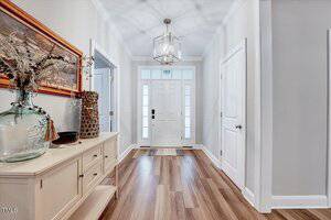 A hallway with wooden flooring features a white sideboard on the left, topped with decorative items and a painting. A white front door with glass panels is at the end. Walls are light-colored, and a modern pendant light hangs from the ceiling.