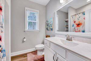 A bathroom with a white vanity and marble countertop, large mirror, toilet, and floral shower curtain. There is a painting with blue flowers on the wall and a window with blinds. A peach-colored rug is on the wooden floor.