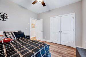A bedroom features a bed with a black and brown checkered bedspread and a pillow shaped like a football. The room has light-colored walls, a ceiling fan, and a closed white double door. There's wall art with letters above the bed.