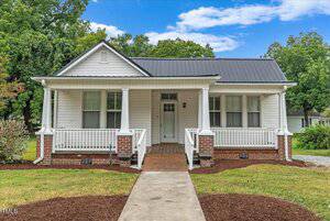 A small white house at 308 Charles Street in Henderson boasts a black metal roof and a covered front porch with white railings and brick accents. It's set against lush green trees and a blue sky. The front yard features a concrete path and well-trimmed grass.