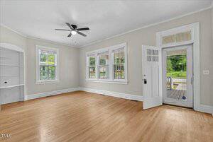 A bright, empty living room features light wood flooring and cream-colored walls. Three windows and a glass door let in natural light. A ceiling fan is centered overhead, and there's a built-in white shelf unit on the left wall.