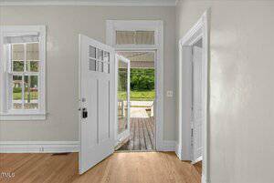 Open white door leading to a wooden porch in a room with light green walls and hardwood floors. There are two windows on the left, and a doorway on the right. Exterior shows greenery and a paved area.