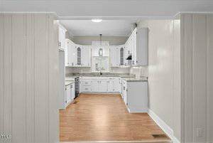 A kitchen with white cabinets and stainless steel appliances. The countertops appear to be granite. The floor is a light wood, and a window above the sink provides natural light. A central light fixture hangs from the ceiling.