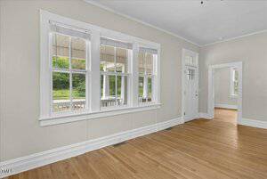 A room with light-colored walls and wooden flooring. It features several large windows with blinds, providing a view of a porch and greenery outside. A doorway opens into an adjacent room with similar decor.
