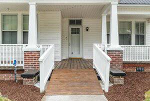The image shows the entrance of a house with a wooden ramp leading to a white door. The house has a white exterior with large windows and brick accents. There is a small light fixture near the door and mulch landscaping on either side of the ramp.