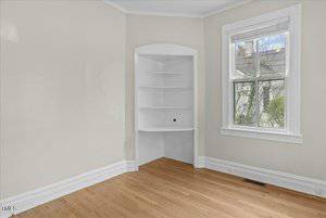 An empty room with light-colored walls and a wooden floor features a small built-in corner shelf. A window with white blinds is on the adjacent wall, allowing natural light. There is a vent near the floor and white baseboards along the perimeter.