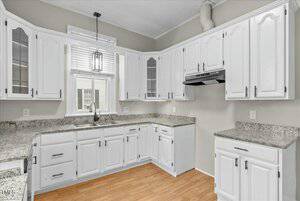 A kitchen featuring white cabinets with black handles, granite countertops, and a single basin sink below a window. An electric stove top with an exhaust hood is present. The floor is light wood, and the walls are painted gray.