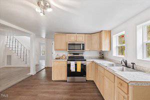 A bright kitchen with wooden cabinets, stainless steel appliances, double sink, and granite countertops. Two windows provide natural light. A staircase and an adjoining room are visible in the background. The floor is dark wood.