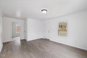 An empty room with light gray wood flooring and white walls. There is a window on the right, a door at the back, and a ceiling light fixture. Another room is visible through an open door on the left.