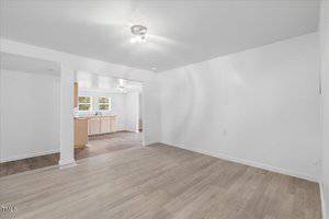 An unfurnished room with light wood flooring and white walls, featuring a single light fixture on the ceiling. An open doorway leads to a kitchen area with wooden cabinets, a window, and visible stainless steel sink.