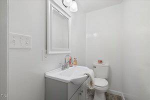 A small bathroom with white walls, featuring a sink with a silver faucet, a mirror above, a hand towel on the sink, a toilet with a candle and jar on the tank, and a ceiling light fixture with two bulbs.