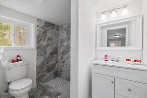 A bathroom featuring a tiled walk-in shower, a toilet beside a window, and a white vanity with a mirror and three overhead lights. The countertop holds soap and red decorative items. The walls are painted white.
