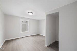 An empty room with light gray walls and wooden flooring. A single ceiling light fixture is on. There's a window with a view of an exterior wall and a small alcove on the right side.