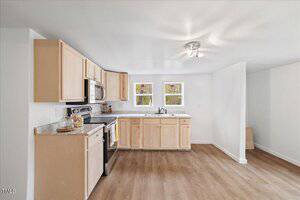 A kitchen with light wooden cabinets and granite countertops. It features a stainless steel oven, microwave, and a sink beneath two windows. The flooring is wood, and the walls are painted white. Ceiling lights illuminate the space.