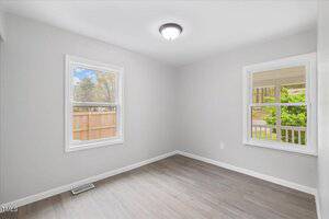 A small, empty room with light gray walls and light wood flooring. Two windows let in natural light; one shows a view of trees and a wooden fence, the other has a view of greenery and part of a porch. A round ceiling light is centered above.