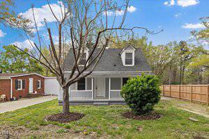 Nestled on 226 1st Street in Henderson, this charming small, two-story house features a gray exterior and a welcoming covered front porch. Two dormer windows grace the roof, while a tree and bush adorn the front yard beside a paved driveway and wooden fence under a blue sky.