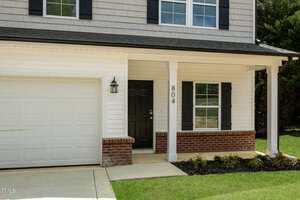 Exterior of a modern two-story house featuring white siding and a brick accent. It has a black front door with a porch area, window with black shutters, and attached garage. The house number "804" is displayed on a pillar. Green lawn surrounds the home.