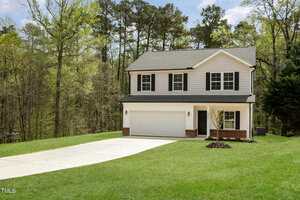 A two-story house with a white and gray facade, black shutters, and a two-car garage. It is situated on a grassy lawn with trees in the background. A small tree is planted in the front yard, and a concrete driveway leads to the garage.