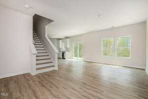 Spacious, empty living area with light wood flooring. Stairs on the left lead to an upper level. Large windows on the right allow natural light. A kitchen in the background features white cabinetry. Pale walls and ceiling contribute to a bright atmosphere.