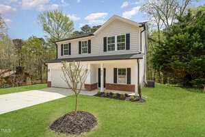 This charming two-story house at 804 E D Street in Butner boasts light-colored siding and dark shutters. It features a welcoming front porch and a single garage, all nestled amid lush greenery, with a small tree gracing the front yard under a partly cloudy sky.