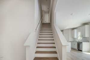 A staircase with wooden steps and white railings leads upward in a well-lit interior. The kitchen is visible to the right, featuring white cabinets and a window. The walls are painted light beige.