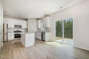 Modern kitchen with white cabinets, stainless steel appliances, and a central island. Light wood flooring extends into an open area with large sliding glass doors leading outside. The kitchen features a gray herringbone tile backsplash.