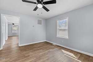 A room with light blue walls and light wood flooring. A ceiling fan with black blades is mounted on the white ceiling. Two windows with blinds let in natural light. An open doorway leads to another room with similar flooring and a ceiling fan.