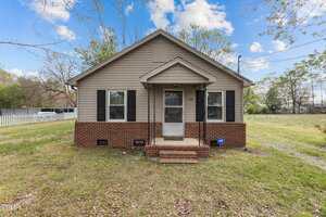 A small single-story house at 128 Warren Avenue in Oxford features beige siding and a brick foundation. The home boasts a gabled roof, black shutters, and a front door with a quaint porch. Lush grass graces the front yard, complemented by a white fence to the left.