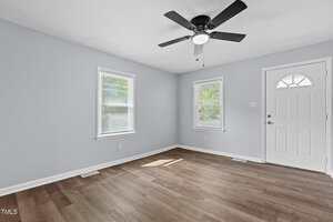 An empty room with light blue walls, two windows with blinds, and a white door. The floor is wood, and a black ceiling fan with a light is mounted on the ceiling. Natural light filters through the windows.