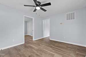 Empty room with pale blue walls, a ceiling fan with black blades, and wooden flooring. Two open doorways lead to adjacent rooms. An air vent and light switch are visible on the walls.
