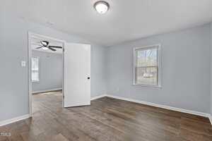 An empty room with light blue walls and wooden flooring, featuring a white door that opens to another room. The room contains two windows with blinds and a ceiling light fixture. The adjacent room has a ceiling fan.