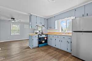 A kitchen with light blue cabinets, wooden countertops, and a stainless steel stove and fridge. The room has a window above the sink, light wood flooring, and a ceiling fan. The adjacent area is visible with similar flooring.