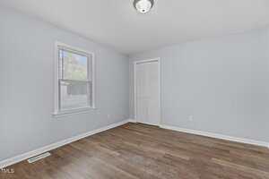Empty, small room with light blue walls, a white ceiling, and laminate wood flooring. A single window with blinds is on the left wall, and a white louvered closet door is on the right. A ceiling light fixture is centered on the ceiling.