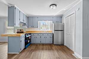 A kitchen with light blue cabinetry and wood countertops. There's a stove with a blue cover, a stainless steel refrigerator, and a window above the sink. The flooring is light wood, and a ceiling light is visible.