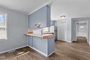 A kitchen with light blue cabinets, wooden countertops, and a white tile backsplash. A white refrigerator is in the corner. Hardwood floors extend into an adjacent hallway. A window with blinds is to the left, and the entrance is visible in the background.