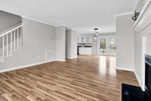 Spacious, unfurnished living area with wood flooring, light gray walls, and a white ceiling. Features include a staircase, fireplace, and open access to the kitchen. Double glass doors in the kitchen lead to an outdoor area.