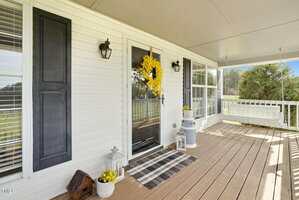 A porch with a wooden floor features a white front door adorned with a yellow floral wreath. There's a black shutter beside the door, a striped doormat, decorative lanterns, and potted plants. A white porch swing is on the right.