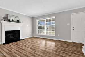 A living room with light gray walls and a white ceiling. It features a white fireplace with a decorative mantel and a large window with blinds. The floor is hardwood. A white door stands on the right side.