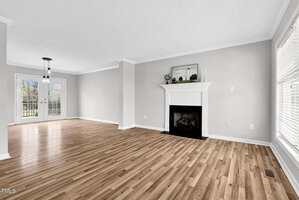 A spacious, empty living room with light wood flooring and light gray walls. A white fireplace is centered on the right wall. Large windows on the right and a hanging light fixture near the back, with double glass doors opening to the outside.