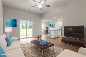 A modern living room with a beige sofa, blue-accented pillows, and a wooden coffee table. A large flat-screen TV is on a low stand. The space has wood flooring, sliding glass doors, and a ceiling fan. The kitchen is visible in the background.