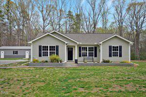 Located at 765 Townsville Landing Road in Henderson, this charming single-story house features light gray siding and a dark gray roof. It boasts a cozy porch with a bench and a detached garage to the left. The front lawn is partially grassy with a few bare patches, framed by a row of trees.