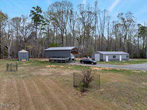 A rural property featuring a carport with a parked black car, a gray shed, and a large gray building, all surrounded by trees and grass. A gravel driveway is visible, and a small fenced area and bare tree are in the foreground.