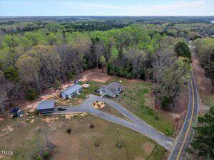 Aerial view of a rural property featuring a house with a circular driveway, surrounded by trees. Nearby are two outbuildings and a separate smaller structure. A road curves along the edge of the property, and expansive greenery surrounds the area.