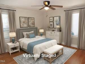 A bedroom features a bed with gray headboard, white bedding, and blue accents. There are matching nightstands with blue lamps, artwork above the bed, and a ceiling fan. A bench sits at the foot of the bed. Text reads "Virtually Staged.