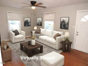 A living room with virtual staging features a white sofa and two white armchairs, a wooden coffee table, and two side tables with lamps. Artworks adorn the walls. A ceiling fan and a "Virtually Staged" label appear in the image.