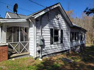 A small, single-story house with white wooden siding, black shutters, and a satellite dish on the roof. There's a screened porch on the left and a wooded area in the background. The yard features some grass and a few trees.