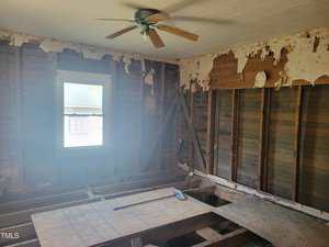 A partially demolished room with exposed wooden wall studs and a missing floor. Peeling wallpaper and an intact ceiling with a fan are visible. Sunlight enters through a window on the left, and debris is scattered on a large wooden board.