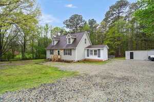 A white, two-story house with a gable roof is set amid a grassy yard. The house has a small wooden porch and gravel driveway. Surrounding trees are green, and there's a gray shed on the right. The sky is clear with a few scattered clouds.