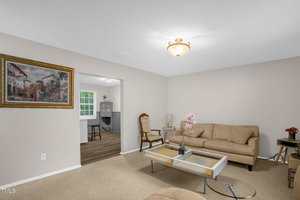 A living room with beige walls and carpet features a tan couch, a glass coffee table, and a wooden chair. A framed painting hangs on the wall. An open doorway leads to a dining area with a round table and a window. Ceiling lights illuminate the space.