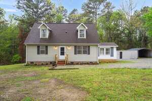 Located at 2603 Brogden Road, this charming beige, two-story house in Creedmoor features a dark roof and is enveloped by lush grass and trees. It boasts a central door with a quaint porch and two dormer windows. A gravel driveway meanders to a white shed under an overcast sky.